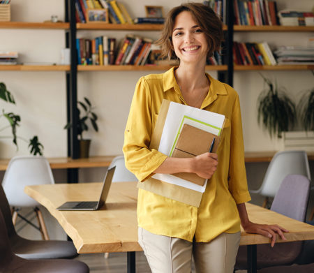 Young cheerful woman in yellow shirt leaning on desk with notepad and papers in hand joyfully looking in camera in modern office